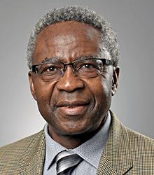 Dr. Funwi Ayuninjam wearing a beige jacket, gray shirt, and gray, black and white tie wearing glasses and smiling at the camera